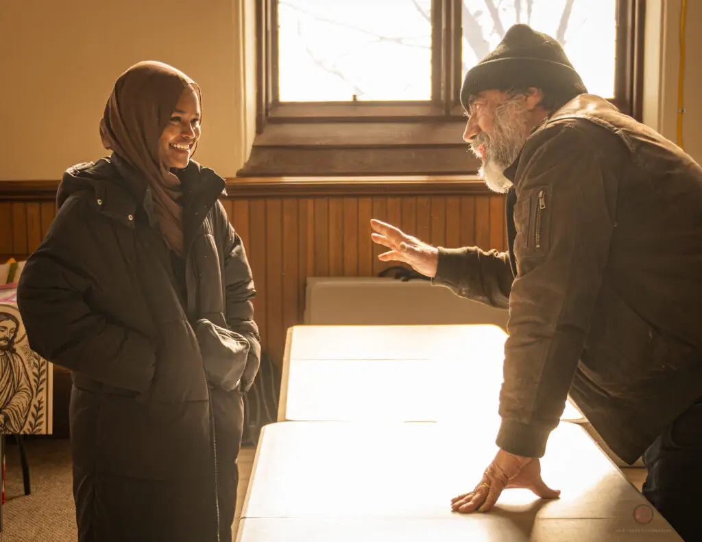 A woman in a hijab stands smiling while a man in a beanie and coat leans on a table, gesturing as they converse in a sunlit room.
