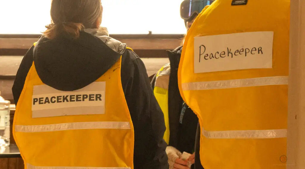 Two people wearing yellow vests labeled “PEACEKEEPER” and “Peacekeeper” stand indoors with their backs facing the camera.