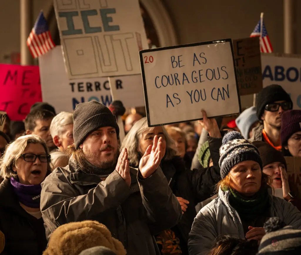 A room full of people with protest signs, hands in the air, and active faces. Signs read "ICE OUT" and "BE AS COURAGEOUS AS YOU CAN"