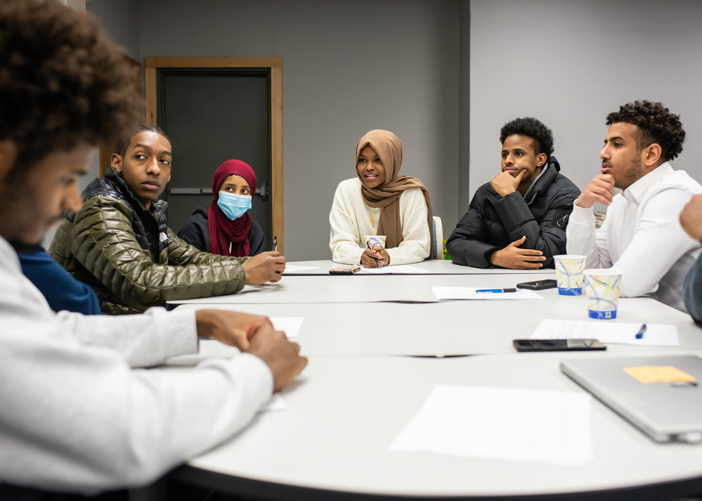 A group of seven people sit around a table in a meeting room, some taking notes and others listening, with papers and cups on the table.