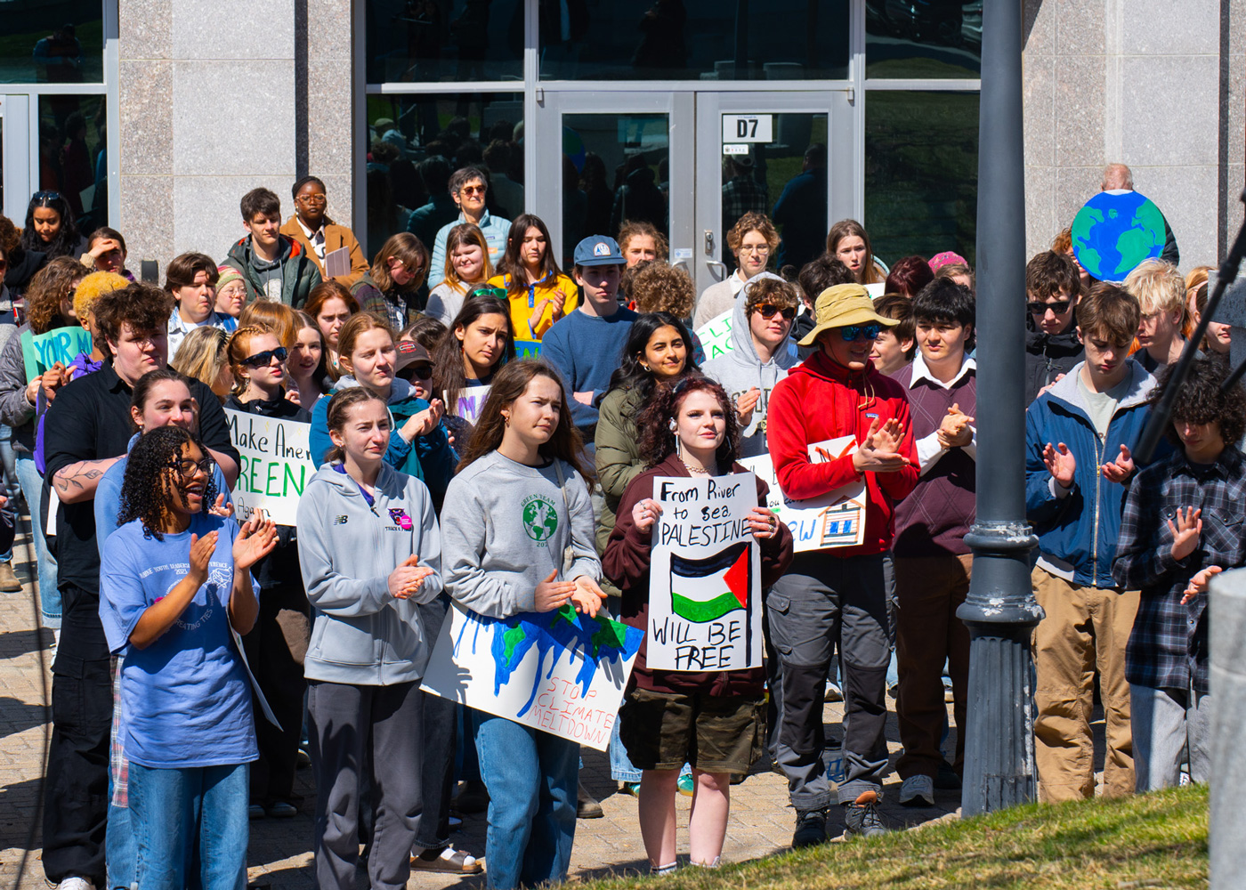 A diverse group of people stand outside a building holding protest signs and clapping during a daytime demonstration.