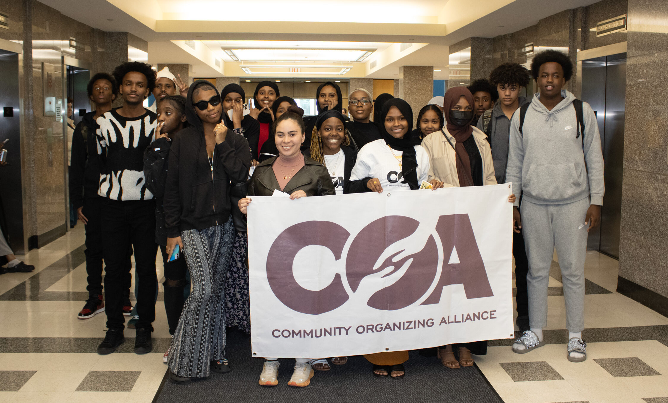 Group of youth smiling for a group photo at the Maine State House, holding a large Community Organizing Alliance banner