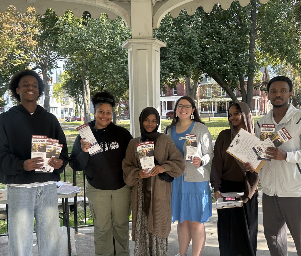 Group of volunteers stand outside on a sunny day, holding clip boards and handouts for Vote No On 1- Save absentee voting in Maine