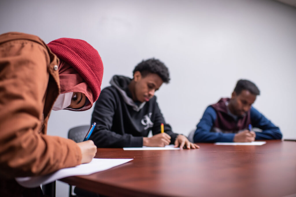 3 youth seated on one side of a table looking down while writing on pieces of paper
