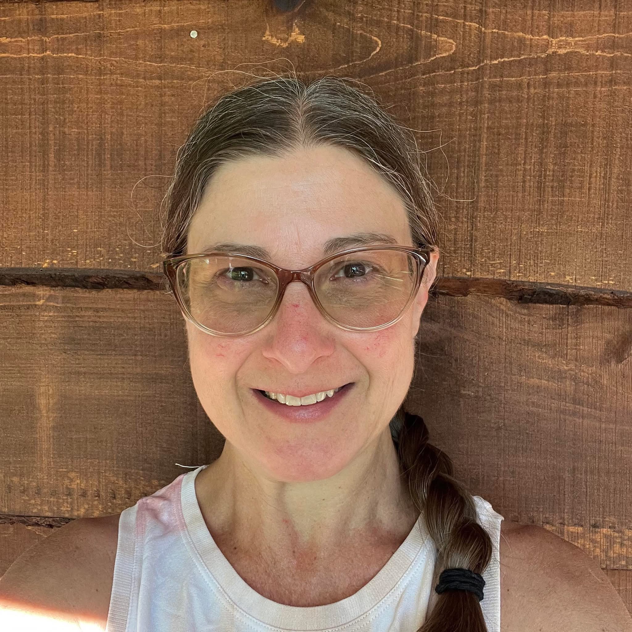 Woman with hair in a side braid with glasses smiling against a wood background