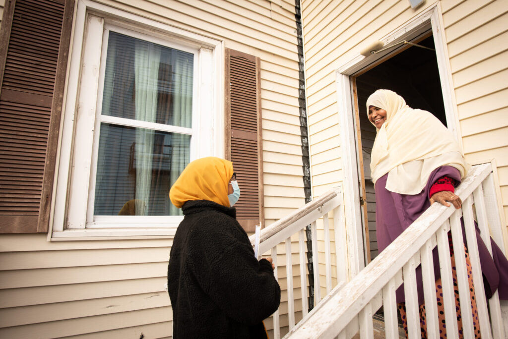 two women are speaking to one another in the front entryway outside of a tan home